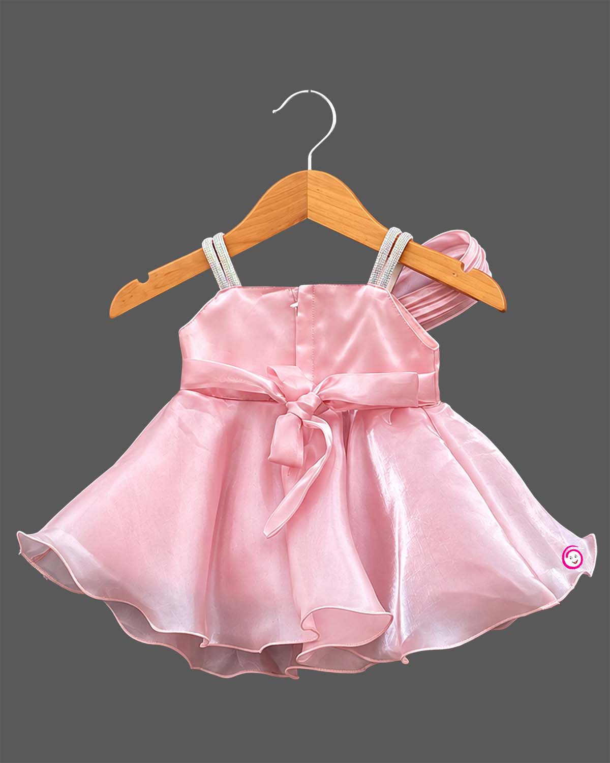 Satin party frock with pleated one-shoulder sash, rhinestone straps, organza overlay skirt, and floral bow applique.