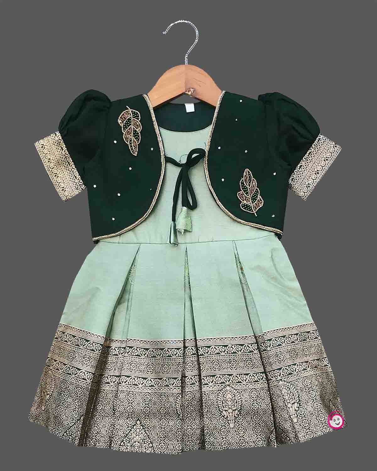 Infant cotton silk frock in pastel mint green and dark bottle green with attached jacket style design, leaf embroidery, front tie-up tassels, and a traditional gold zari border