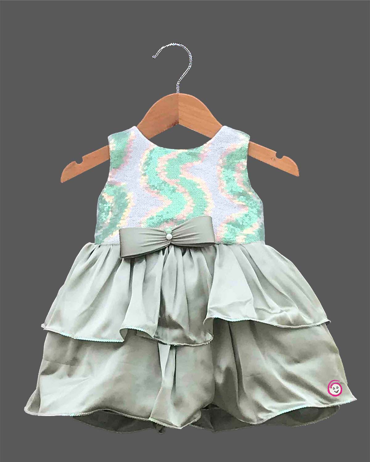 Little girl's party wear frock featuring a pastel green, pink, and white sequined sleeveless bodice and a tiered sage green satin skirt with a central bow on a wooden hanger against a grey background.