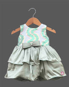 Little girl's party wear frock featuring a pastel green, pink, and white sequined sleeveless bodice and a tiered sage green satin skirt with a central bow on a wooden hanger against a grey background.