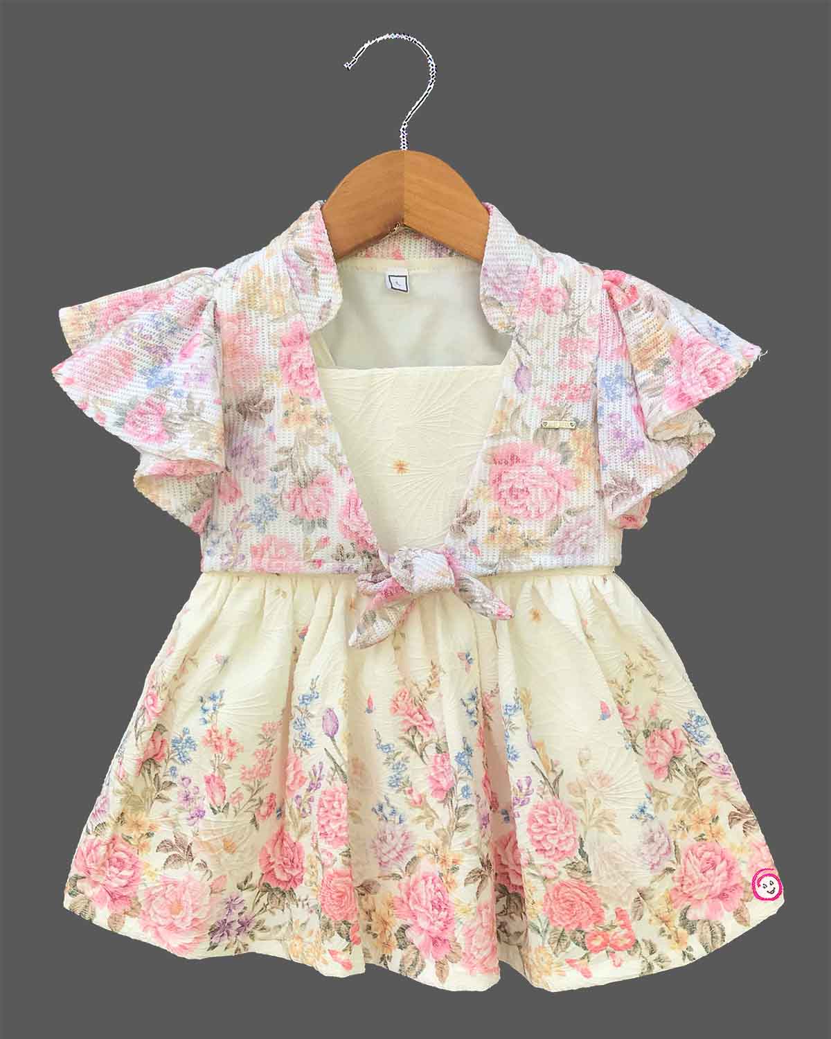 Elegant floral printed baby girl frock with layered flutter sleeves, a front knot accent, and a textured flared skirt displayed on a wooden hanger.