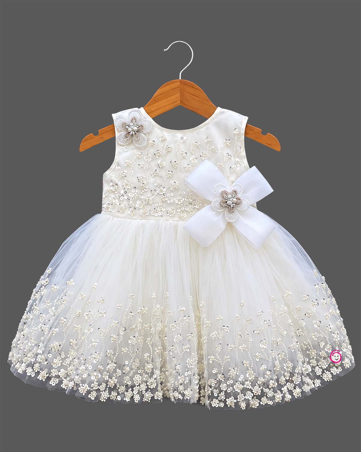 Sleeveless net frock with sequin bodice, waist bow motif, applique shoulder flower, and embroidered hem.