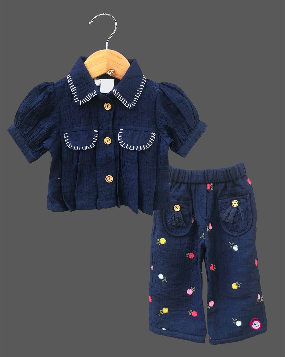Stylish two-piece set for baby girls featuring a collared short-sleeve top with contrast stitch trim and matching embroidered pants with tassel details on a hanger.