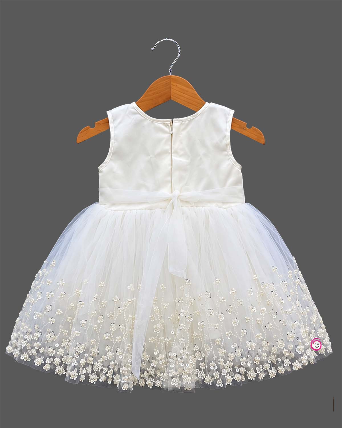 Sleeveless net frock with sequin bodice, waist bow motif, applique shoulder flower, and embroidered hem.