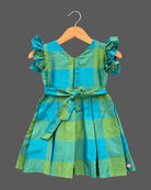 Girls checked silk cotton frock featuring V-neck, ruffle flutter sleeves, and pleated skirt with elephant zari border.