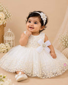 Sleeveless net frock with sequin bodice, waist bow motif, applique shoulder flower, and embroidered hem.