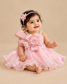 Satin party frock with pleated one-shoulder sash, rhinestone straps, organza overlay skirt, and floral bow applique.