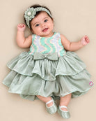 Little girl's party wear frock featuring a pastel green, pink, and white sequined sleeveless bodice and a tiered sage green satin skirt with a central bow on a wooden hanger against a grey background.